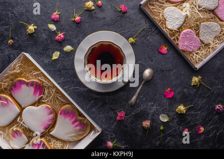St Valentines Tag flach mit Tasse Tee, verglaste herzförmige Plätzchen und dekorative Blumen auf dunklen Tischplatte Stockfoto