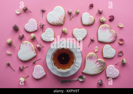St Valentines Tag Anordnung der Tasse Tee, verglaste herzförmige Plätzchen und dekorative Blumen isoliert auf Rosa Stockfoto