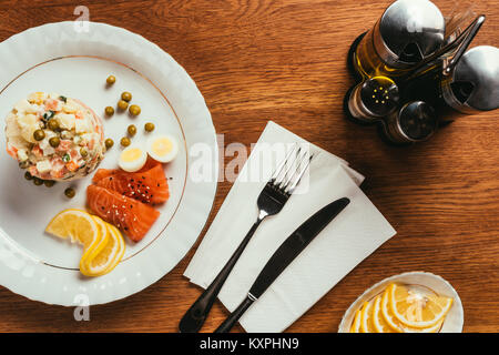 Russischer Salat auf Teller mit verstreuten Erbsen, gekochten Eiern und Fisch Schichten über Tabelle mit Gabel und Messer auf Serviette Stockfoto