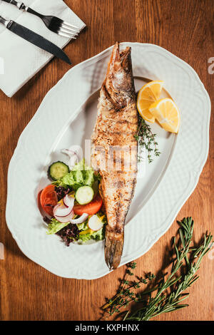 Gebratener Fisch auf weiße Platte mit Gemüse und Orangenscheiben über Holz- Oberfläche mit Gabel und Messer auf Serviette Stockfoto