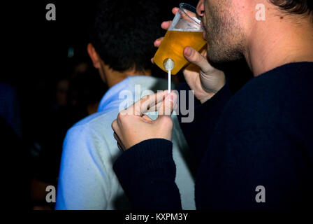 Ein 7/8-Ansicht von einem jungen Mann in seinen Zwanzigern trinken Bier aus einem kleinen Plastikbecher während holidng einen Lutscher in einem Nachtclub Stockfoto