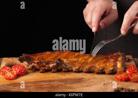 Der Koch schneidet sie mit einem scharfen Messer bereit Rippchen essen, liegend auf einem alten Holztisch. Ein Mann bereitet einen Snack zum Bier auf einem schwarzen Hintergrund mit Kopie Raum Stockfoto
