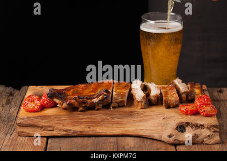 Schweinerippchen in Barbecue Sauce und Honig gebackene Tomaten auf dem alten Holztisch. Fleisch und bier auf schwarzen Hintergrund mit kopieren. Im Hintergrund schüttete das Bier in das Glas Stockfoto