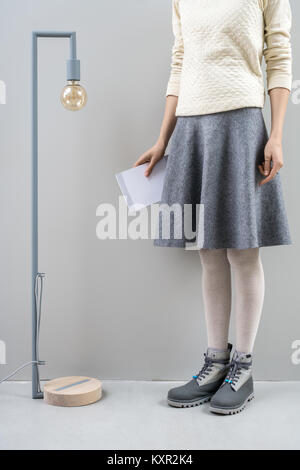 Charmante Mädchen steht neben dem langen Lampe mit Edison die Glühbirne auf die graue Wand Hintergrund im Studio. Sie trägt einen beigefarbenen Pullover, Grauer Rock, Strumpfhose Stockfoto