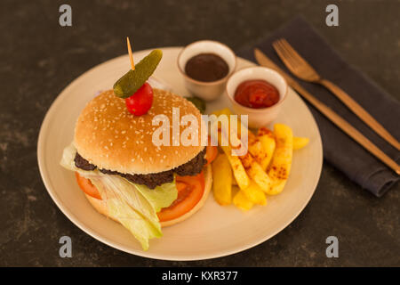 Hamburger mit Pommes Frites, Tomaten und Barbeque Sauce in beige Keramikplatte auf Schwarz rustikalen Tischplatte mit Kupfer Besteck gelegt. Stockfoto