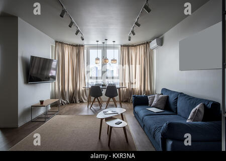 Elegante und moderne Einrichtung mit weißen Wänden und einem Parkettboden mit einem Teppich auf dem Boden. Es ist ein blaues Sofa mit Kissen, Holztische und Stühle, TV, stand Stockfoto
