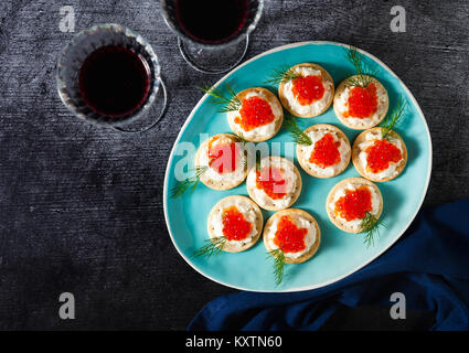 Festliche Portion roter Kaviar Sandwiches auf einem blauen Schild mit einem dunklen Stein Hintergrund und zwei Gläser Rotwein Stockfoto