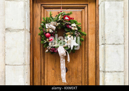 Vorderansicht eines traditionellen Weihnachten Krone aus immergrünen Zweigen, roten Weihnachtskugeln, Holly Blätter und Beeren auf einer hölzernen Tür hing mit Stockfoto