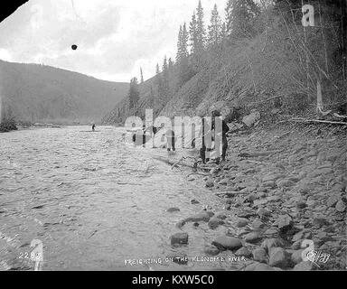 Foto aus der Zeit um 1898, das vier Männer zeigt, die während des Klondike Gold Rush ein kleines Boot am Ufer des Klondike River im Yukon-Territorium ziehen und so das Leben und die körperliche Arbeit der damaligen Zeit einfangen. Stockfoto