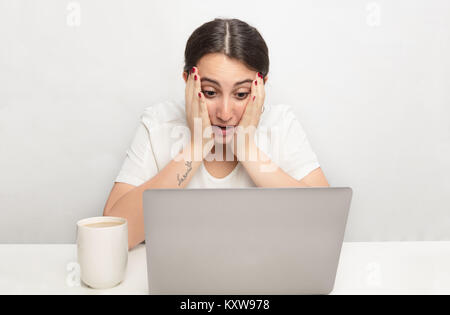 Junge Frau an einem Laptop in Unglauben und Schock ihr Gesicht ruhen auf ihre Hände, als sie auf den Bildschirm starrt starrte Stockfoto