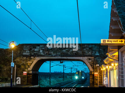 Drem Bahnhof, East Lothian, Schottland, Großbritannien, in der Abenddämmerung mit Licht auf ScotRail Zug und nähert sich unter einer Brücke Stockfoto