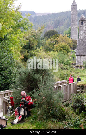 Man Straßenmusik in der schottischen Soldaten Outfit sitzend durch seine Rüstung und spielt Dudelsack am Eingang zur Klosteranlage von Glendalough Valley gekleidet Stockfoto