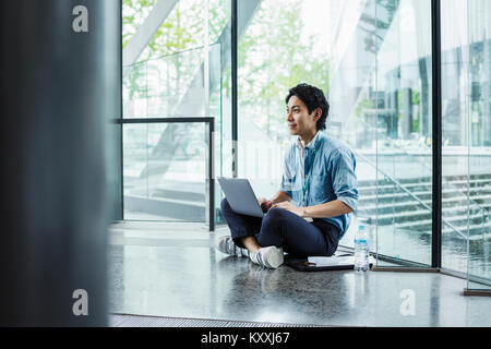 Unternehmer tragen blaue T-Shirt sitzen auf dem Boden im Innenbereich, lehnte sich gegen die Glaswand, Arbeiten am Laptop. Stockfoto