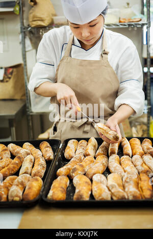 Frau mit Kochmütze und Schürze in einer Bäckerei arbeiten, Schneiden frisch gebackenen Brötchen auf großen Tabletts. Stockfoto