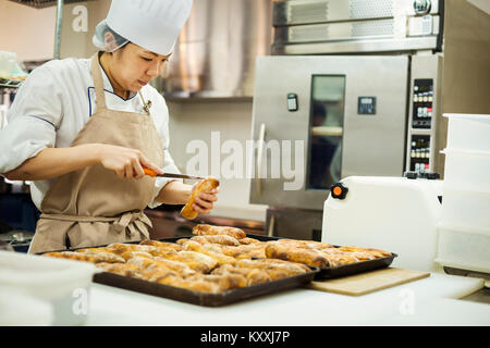 Frau mit Kochmütze und Schürze in einer Bäckerei arbeiten, Schneiden frisch gebackenen Brötchen auf großen Tabletts. Stockfoto