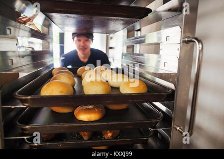 Mann, der in einer Bäckerei arbeitet, indem große Fächer mit frisch gebackenen Brötchen auf einem Trolley. Stockfoto