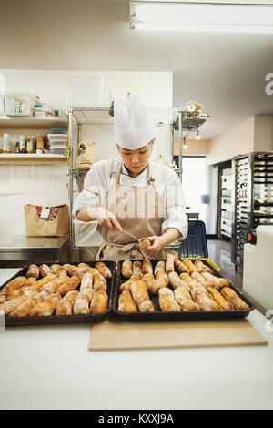 Frau mit Kochmütze und Schürze in einer Bäckerei arbeiten, Vorbereitung frisch gebackenen Brötchen auf großen Tabletts. Stockfoto