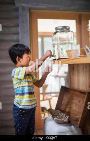 Junge mit schwarzen Haar zu tragen gestreifte T-shirt Füllung Glas mit Wasser aus dem Wasserspender. Stockfoto