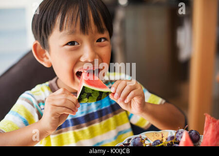 Der Junge mit den schwarzen Haaren tragen gestreifte T-Shirt essen ein Stück Wassermelone. Stockfoto