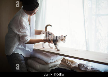 Frau in Innenräumen, holding Calico Katze mit weißen, schwarzen und braunen Fell Wandern auf einem Regal. Stockfoto