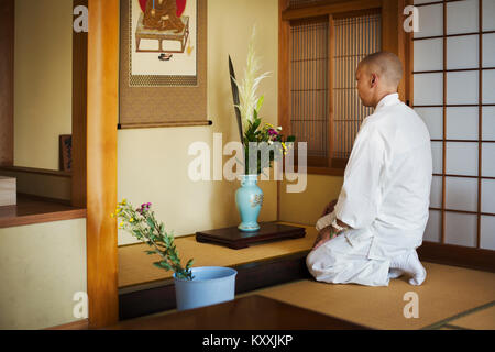 Seitenansicht des buddhistischen Mönch mit rasiertem Kopf tragen weiße Robe kniend vor Vase mit Blumen. Stockfoto