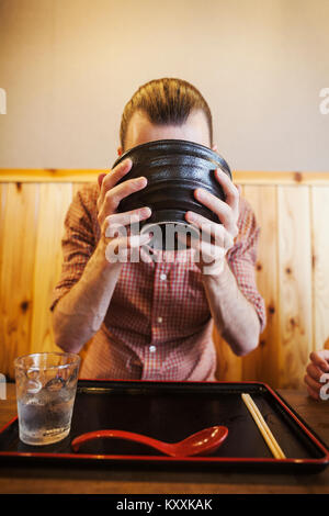 Ein westlicher Mensch in eine Nudel Restaurant, mit einer Nudel Schüssel es Heben mit zwei Händen daraus zu trinken. Stockfoto