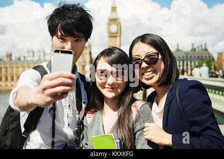 Lächelnd Mann und zwei Frauen mit schwarzen Haaren unter selfie mit Smartphone, stehend auf die Westminster Bridge über die Themse, London, mit den Häusern Stockfoto