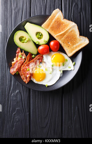 Köstliches Frühstück mit Eiern mit knusprigem Speck, Avocado, Toast und Tomaten close-up auf schwarzem Teller auf dem Tisch. Vertikal oben Ansicht von oben Stockfoto
