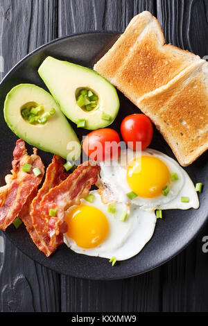 Herzhaftes Frühstück: Spiegeleier mit Speck, Avocado, Toast und Tomaten close-up auf einem Teller auf dem Tisch. Vertikal oben Ansicht von oben Stockfoto