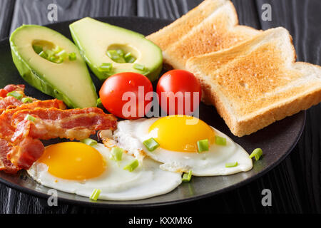 Gesunde traditionelle Frühstück mit Spiegelei mit Speck, frischen Avocado, Toast und Tomaten closeup auf schwarzem Teller auf dem Tisch. Horizontale Stockfoto