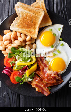 Englisch, hausgemachtes Frühstück: Spiegeleier mit Speck, Bohnen, Toast und frischem Gemüse auf einem Teller auf den Tisch. Vertikal oben Ansicht von oben Stockfoto