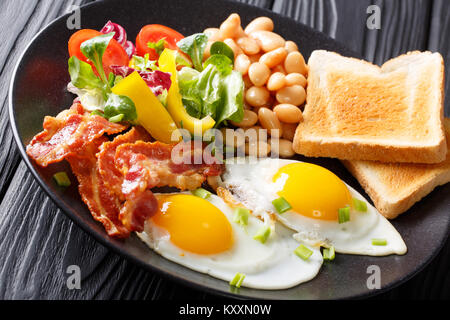 Englisch gesundes Frühstück: Zwei Spiegeleier mit Speck, Bohnen, Toast und Salat auf schwarzem Teller auf dem Tisch. Horizontale Stockfoto