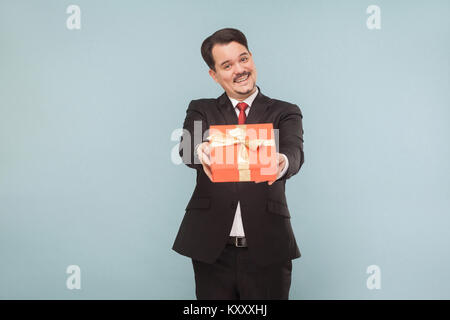 Glücklicher Mann im schwarzen Anzug und mit roter Geschenkbox. Studio shot, auf hellblauem Hintergrund isoliert Stockfoto