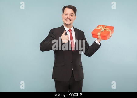 Glücklicher Mann im schwarzen Anzug mit roten Geschenkkarton mit Daumen nach oben. Studio shot, auf hellblauem Hintergrund isoliert Stockfoto