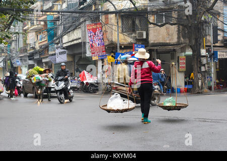 Hanoi, Vietnam - 13. Dezember 2017. Eine Straße Verkäufer Spaziergänge über eine befahrene Kreuzung mit ihrer Schulter-/Tragetasche Pole mit 2 Körben Stockfoto