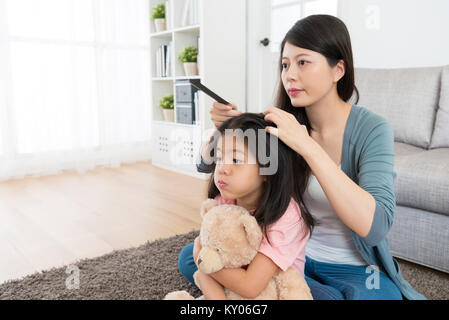 Junge süsse kleine Mädchen, dass Teddy Spielzeug auf dem Boden sitzen in Morgen und Gesicht Fratze im Spiegel zu betrachten, wenn die Mutter die Haare kämmen. Stockfoto