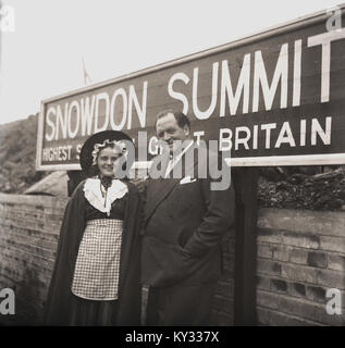 1950, historische, Abbildung, die den bekannten und viel beachteten BBC-Reporter Richard Dimbleby und ein junges Mädchen in die Welsh National Kostüm mit Schürze und Haube stehen zusammen auf dem Gipfel des Mont Snowdon in Wales, der höchste Gipfel in der Britischen Inseln. Stockfoto