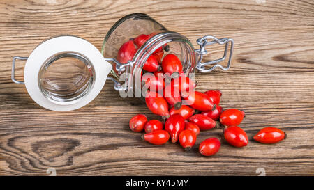 Verschüttete rote Hagebutten auf Holz Tisch. Nahaufnahme der Lüge jar und Arzneimittel Früchte von Wild Rose auf den Jahrgang Hintergrund. Stockfoto