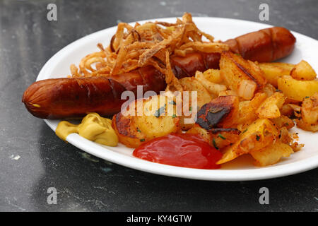 Nahaufnahme, Teil von einem grossen Bratwurst mit hausgemachtem geröstete Kartoffeln, gebratenen Zwiebeln, Ketchup und Senf auf weiße Platte über Grau, Stockfoto