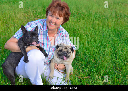 Fröhliche Frau, Mutter, Hausfrau, halten zwei Haustiere, Hunde. Schwarz, aktiv, neugierig Französische Bulldogge, beige, Rehbraun, dicke Mops. ältere Frau schaut zu. Stockfoto