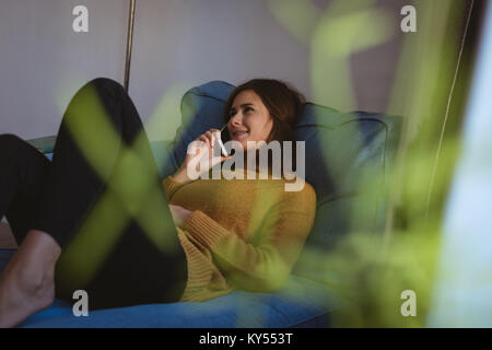 Lächelnde junge Frau liegt auf einem Sofa im Gespräch über Ihr Mobiltelefon im Wohnzimmer Stockfoto