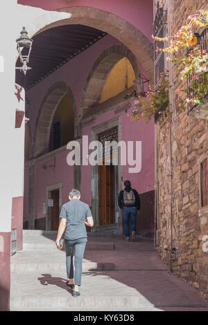 Fußgänger gehen auf eine hügelige Stein Gehweg zu einem großen gewölbten Eintrag, mit einer Laterne und andere architektonische Details, in Guanajuato, Mexiko Stockfoto