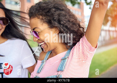 Attraktive junge Frau Gesang zur Musik, während Ohrhörer und gehen mit ihrer Freundin auf der Straße im Sommer Stockfoto
