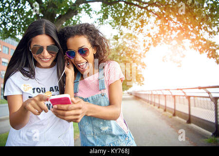 Zwei unbeschwerte junge Freundinnen lächelnd und gehen hinunter eine Promenade im Sommer Musik zusammen auf ein Paar Kopfhörer Stockfoto