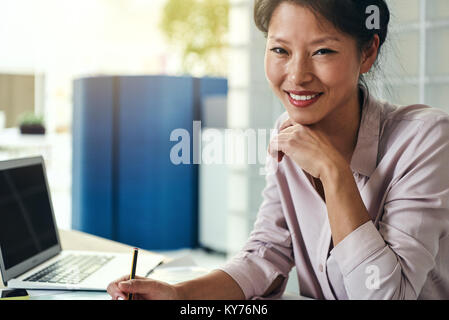 Selbstbewussten jungen asiatischen Geschäftsfrau, lächeln, wenn sie alleine an einem Tisch in einem modernen Büro arbeiten Stockfoto