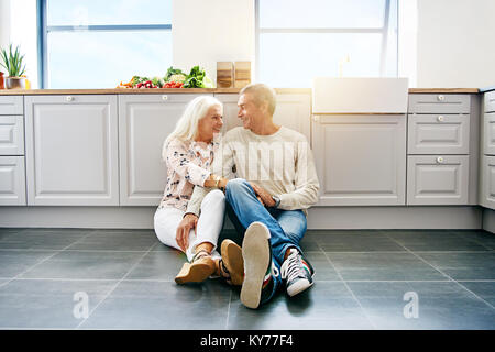 Herzlichen senior Paar lächelnd und an einander, während sie zusammen auf dem Boden ihrer hellen Küche zu Hause sitzen Stockfoto