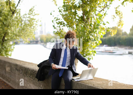 1001 Manager arbeiten mit Laptop in der Nähe von Fluss und Argumentieren mit Partner von Smartphone. Hübscher junger Mann im schwarzen Anzug mit blauer Krawatte suchen Stockfoto