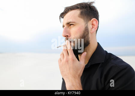 Geograph männliche Raucher, junger Mann kommen, um am Meer zu erforschen. Kerl mit kurzen Haar steht auf Sand anzeigen Papiere in Schwarz fo Bärtigen Stockfoto