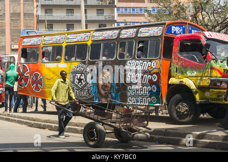 Ein Mann schiebt seine Hand Warenkorb auf der Straße hinter einem hell eingerichtete Bus auf dem Bürgersteig, Nairobi, Kenia, Ostafrika Stockfoto