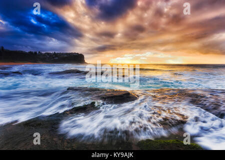 Dunkle Sonnenaufgang über bungan Strand von Sydney Northern Beaches Pazifikküste mit stürmischen Wellen über rollende Felsen unter bewölktem Himmel mit steigenden zu Sandstein Stockfoto